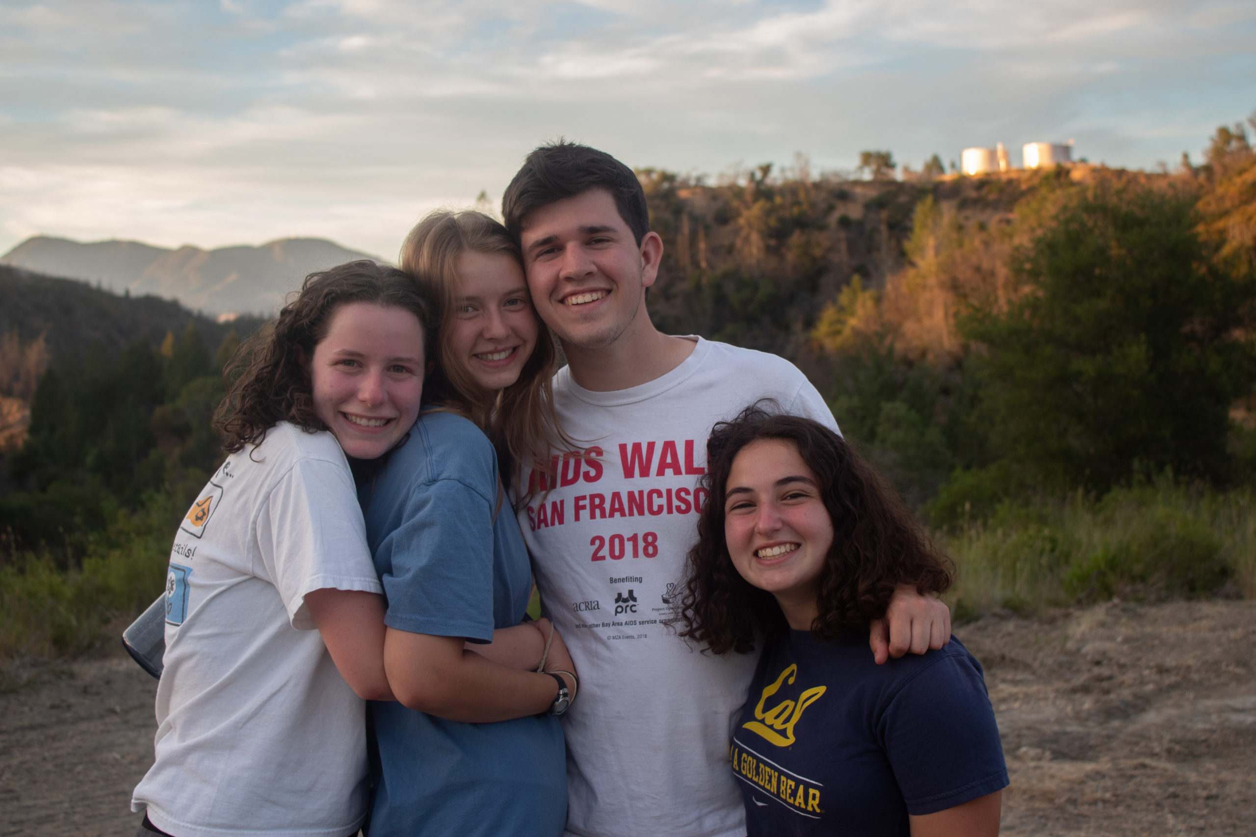 Group of four college-aged camp counselors hugging with a sunset in the background at summer camp.